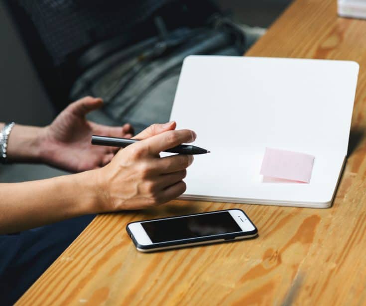 A man taking notes in a notebook. A smart phone sits next to it.