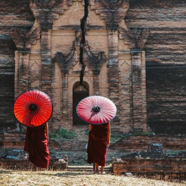 Two individuals in traditional attire hold red umbrellas while standing in front of an ancient, cracked structure made of brick, embodying the kind of dedication and passion we seek. Junior Account Manager required to join our team.