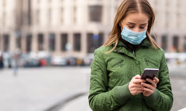 woman in a green jacket wearing a mask and looking at her phone on a city street