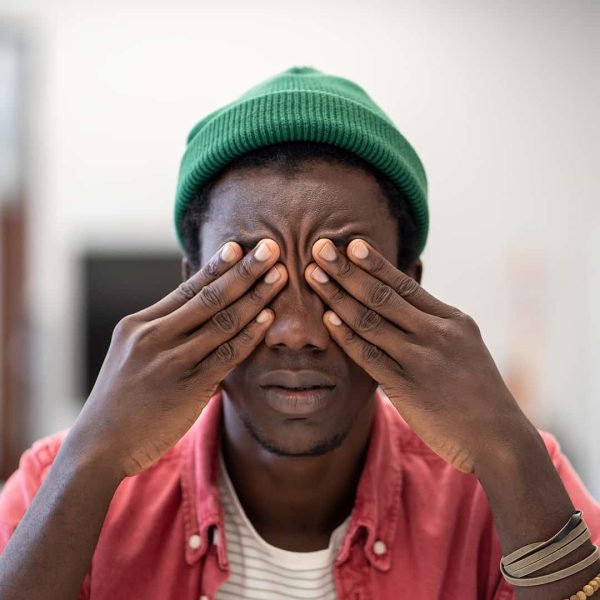 A person wearing a green beanie and red shirt covers their eyes with both hands, appearing tired or stressed, perhaps from the search for a Junior Account Manager required to join our team.