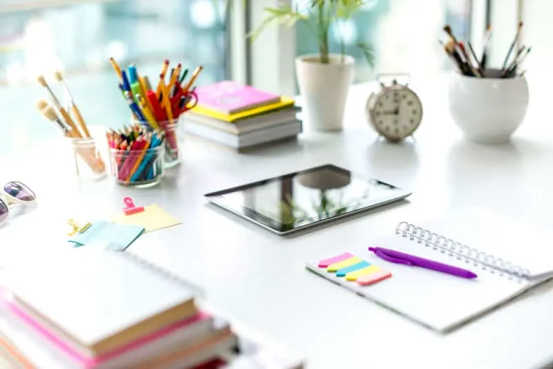 A desk with a planner, notebooks, tablet, and cups of writing utensils.